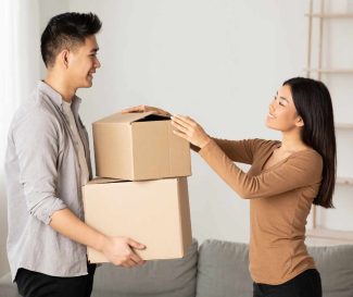 Smiling couple carrying cardboard boxes in their living room, preparing for a move. Their move was made stress free when they choose one of the best movers in Singapore.